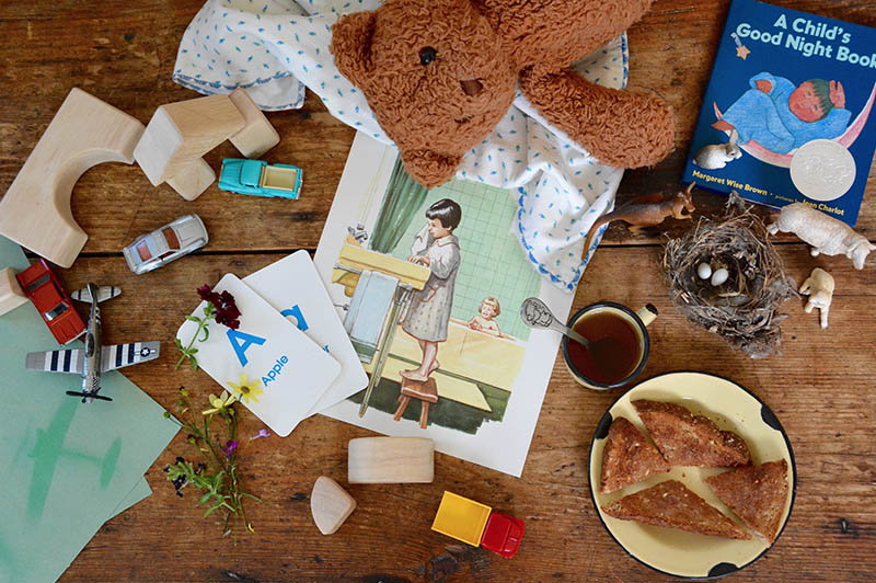 Looking down on a wooden table, the small, blue Caldecott Award-winning picture book, A Child's Good Night Book by Margaret Wise Brown is in the upper right with a mess of preschool arts and crafts projects and a snack of buttered toast and cambric tea strewn about.
