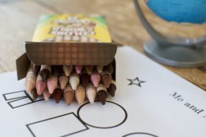 An opened box of Crayola's Colors of the World crayons are set on a coloring page. A small globe is next to the crayons on the wooden table.