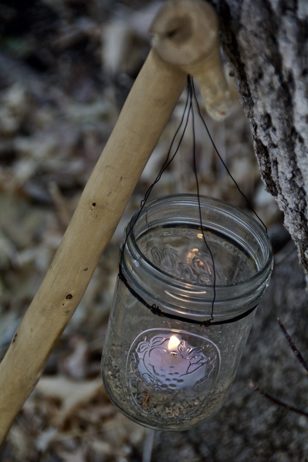 mason jar candle hanging from a wooden cane leaning against a tree trunk. Autumn leaves are on the ground.