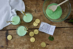 St. Patrick's Day display on wooden table. Green, pistachio pudding served in individual clear glasses with ornate gold spoons. Gold foil chocolate coins and Lucky Leprechaun note displayed on table.