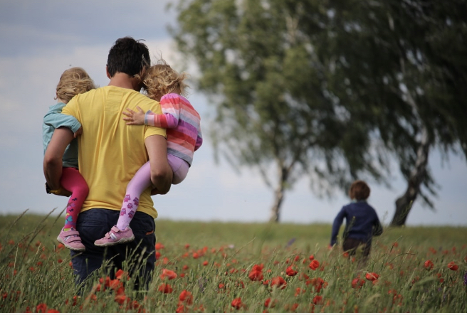 a dad carrying two little children in a field of red flowers. A son is running ahead of everyone.