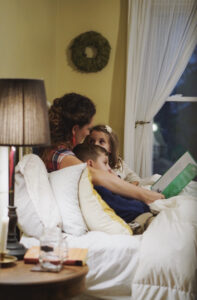 As part of their Children's Hour - a nighttime routine, a mother is reading a book to her young daughter and son. They are sitting on a bed together in a yellow room with a lamp lit on the bedside table and a curtained window next to them.
