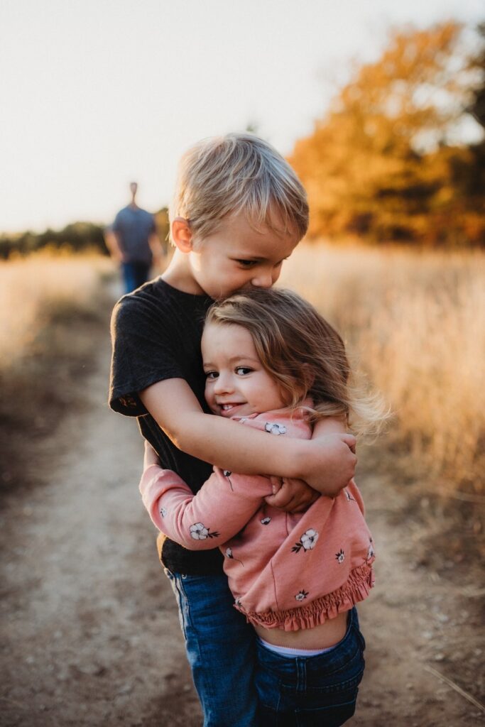 A young brother and sister hug outside on a hike enjoying an autumn activity at home with their dad.