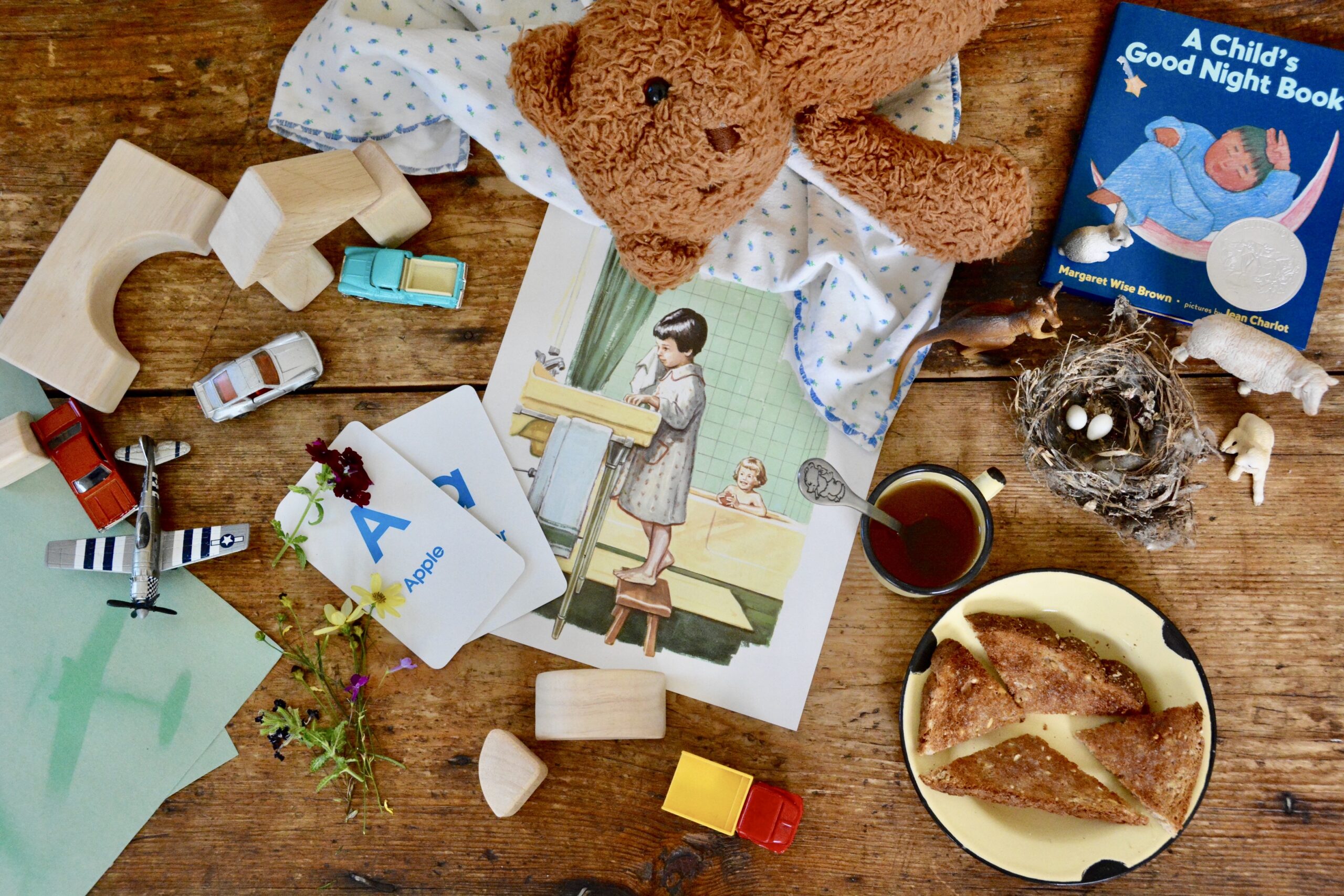 a messy collection of materials on a wooden table used for a preschool lesson including a teddy bear, Aa flash cards, small cars, wooden blocks and a snack of tea and toast.