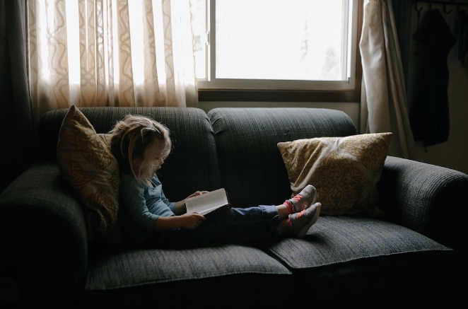 A young child sitting on a gray couch next to a window and reading.