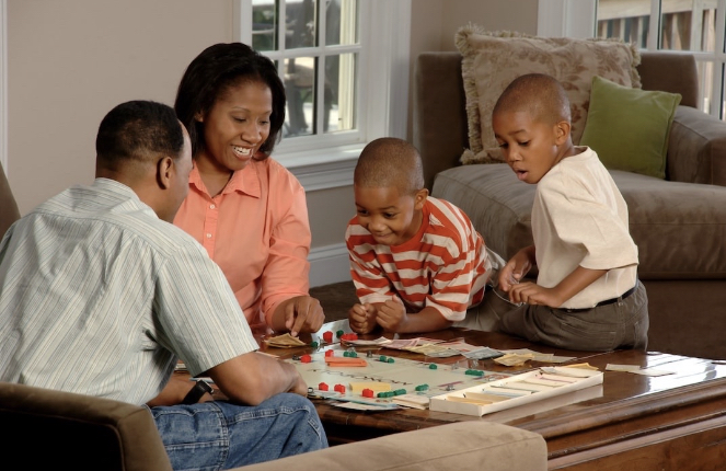 A family consisting of two elementary school age boys and their mom and dad are playing monopoly together in their family room.