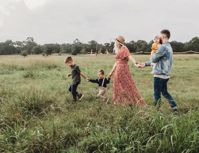 A young family holding hands and walking outside in a field. The mother is dressed in a long red dress with a straw hat. The dad is holding a little baby girl and the toddler and a young boy are leading the group.