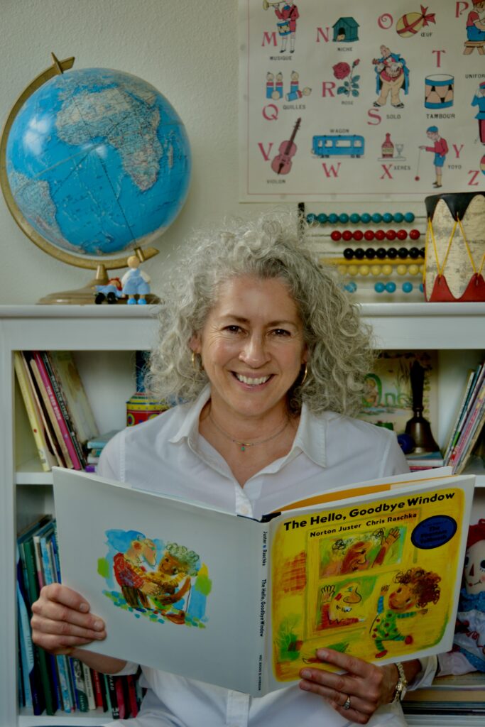 A gray haired teacher reading a picture book in front of a bookcase with a globe and vintage toys set on top.