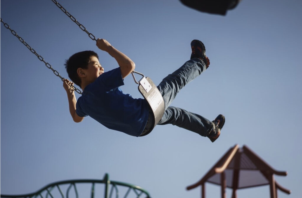 preschooler boy wearing a blue t-shirt and jeans is swinging high on a swing