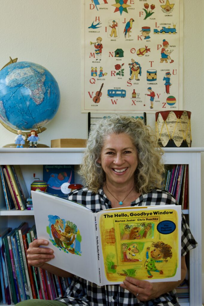 a woman with a black and white checkered shirt is reading a picture book infant of a bookcase full of preschooler toys and books.