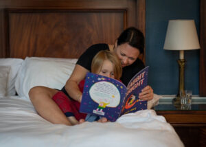A brunette mother and young girl are sitting on a bed and reading a blue picture book together.