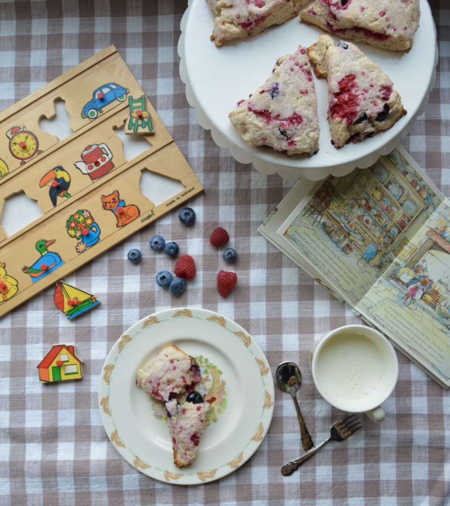 A preschool child's snack of a scone and milk is set on a beige and white checkered tablecloth with berries, a puzzle and picture book.