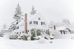 A white double story home with a red brick chimney is enveloped in snow under a white, snowy sky exuding a feeling of shelter and deep winter joy at home.