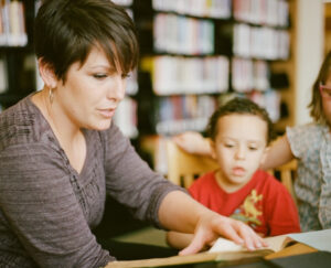 A pretty mother with dark hair and a cropped hairstyle wearing a brown shirt is reading children's poetry to her two young children, son and daughter, in a public library.