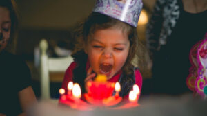 fun, happy and excited preschooler blowing out candles on her birthday cake wearing a hat