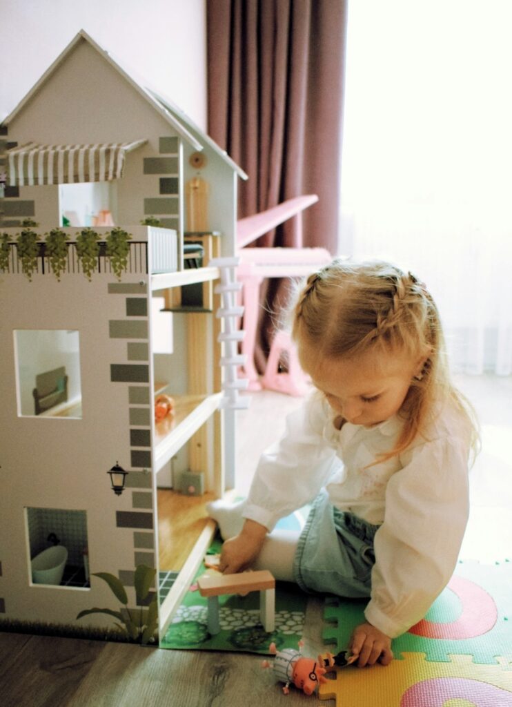 blonde little preschooler girl in braids sitting on the floor playing with her dollhouse